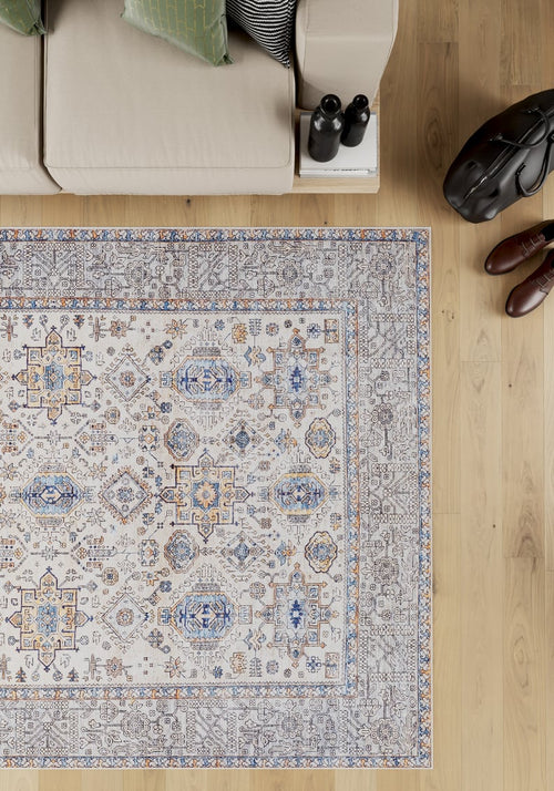 Load image 1 in gallery view. The Nexa Bordered Beige Washable Rug (LCC3009), featuring blue, gold, and grey patterns, lies on a light wood floor near a beige sofa with green pillows. Brown shoes and a black bag are placed next to the sofa.