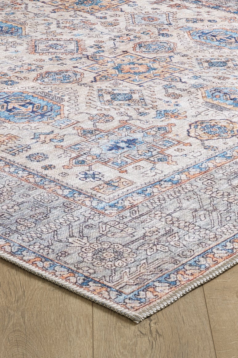 A close-up of the Nexa Bordered Beige Washable Rug - LCC3009 (Custom Size) shows its blue, beige, and rust geometric pattern with detailed borders on a light wood floor. This washable rug has a slightly faded vintage look, ideal for high-traffic areas.