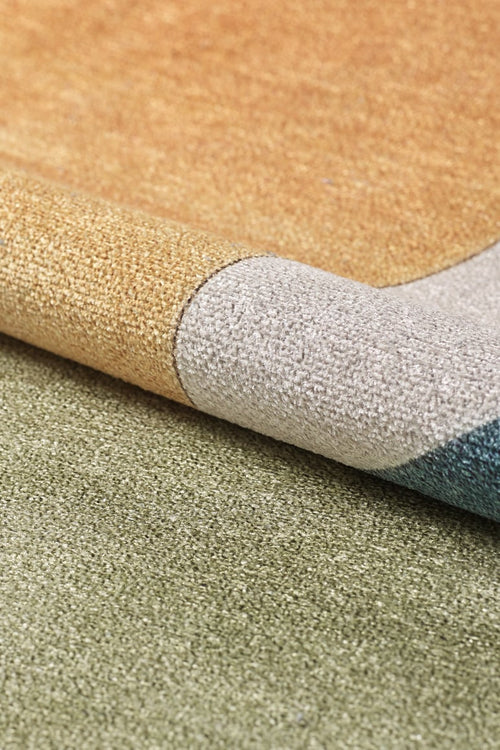 Load image 1 in gallery view. The Nexus Geometric Contemporary Washable Rug - MDJ006, in gold, gray, blue, and green tones, is shown slightly folded on a green carpet—an ideal choice for high-traffic spaces.