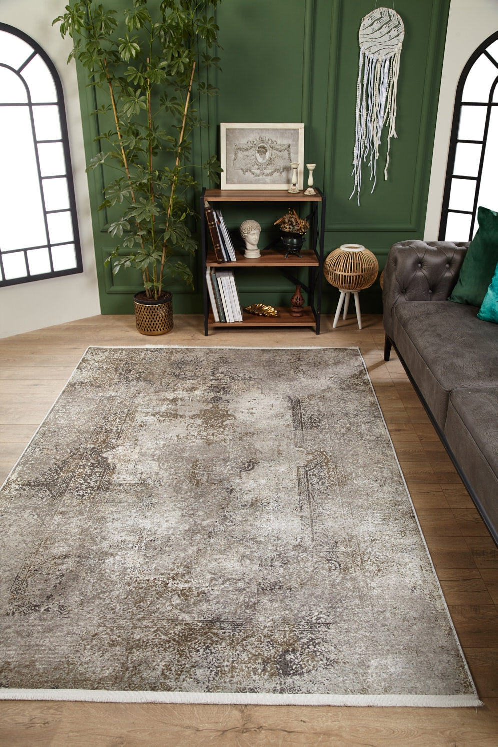 A modern living room with a Noble Insignia - Washable Rug - JR1830 (Custom Size) in distressed beige, gray tufted sofa, green accent wall, bookshelf with decor, tall plant, and arched windows. Ideal for high-traffic areas.
