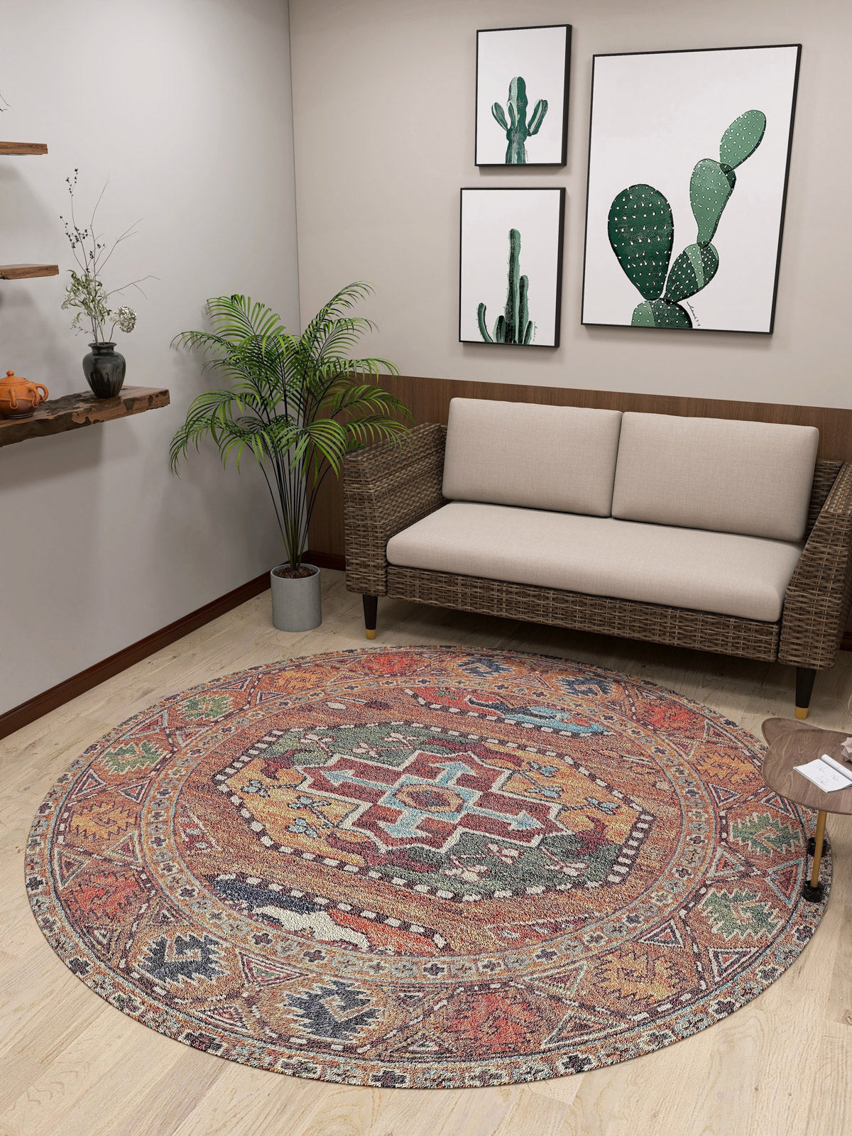 A small room features a beige sofa, a plant, and the Nomadic Oasis - Washable Rug - JR5086. Three framed cactus prints hang above the sofa near a wooden shelf decorated with small plants and pottery.