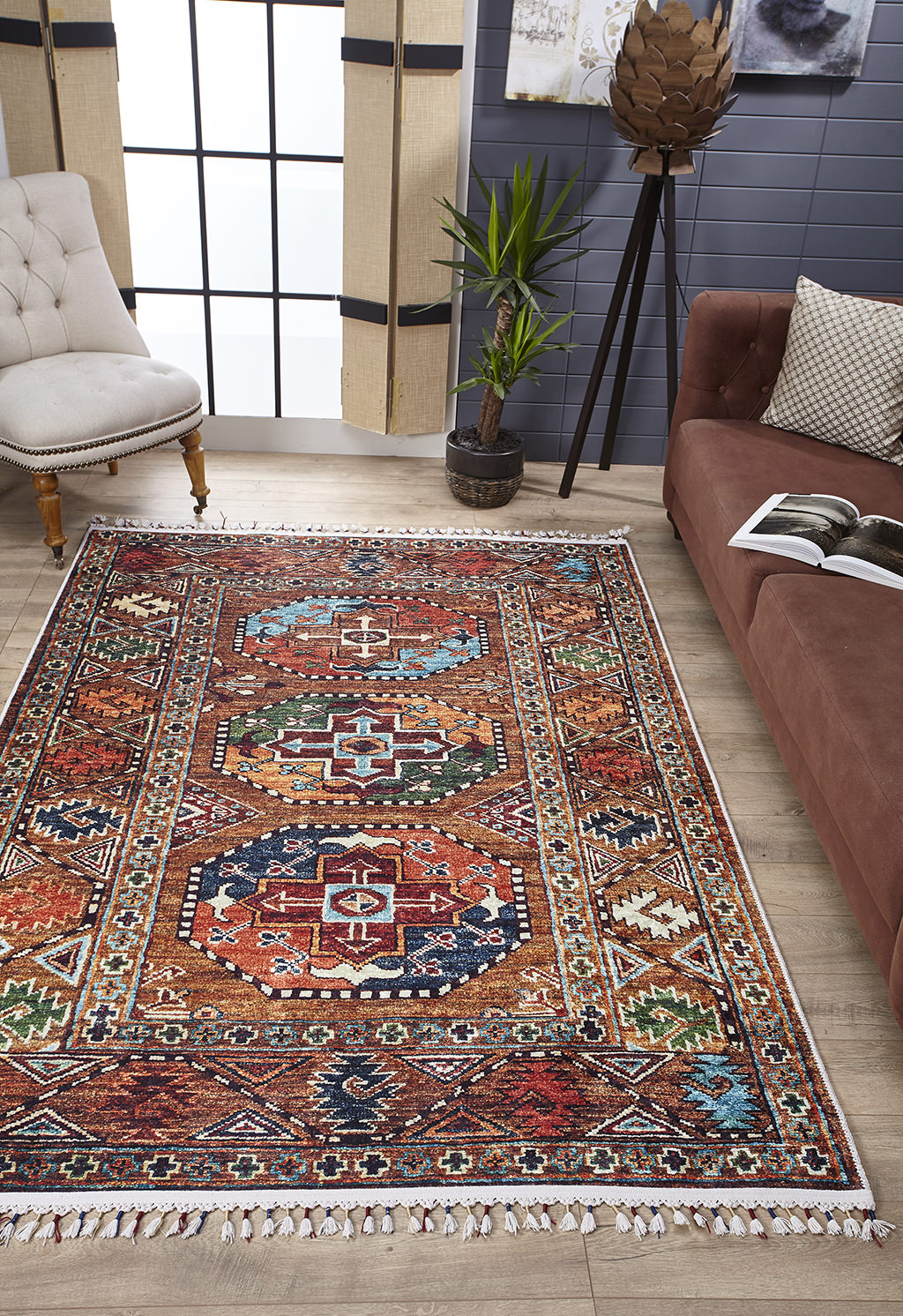A living room with the Nomadic Oasis - Washable Rug - JR5086 (Custom Size), featuring colorful geometric designs—ideal for high-traffic areas—plus a brown sofa with an open magazine, a white tufted chair, tall potted plant, and sheer-curtained windows.