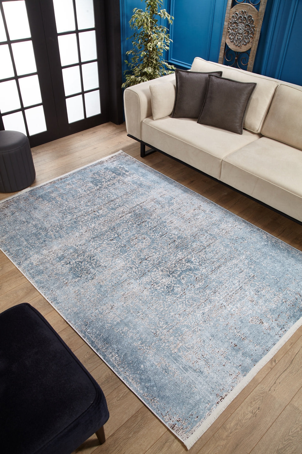 A modern living room featuring a cream sofa, dark cushions, the Old World Charm - Washable Rug - JR1606 (Custom Size) in blue for high-traffic wooden floors, a potted plant, blue accent walls, and black-framed glass doors.
