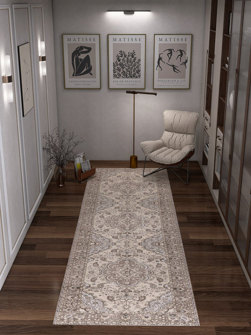 Load image 1 in gallery view. A hallway with dark wooden floors features the Opulent Mirage - Washable Rug - JRN1008 in a subtle medallion design, alongside a cushioned chair, floor lamp, glass vase with branches, framed Matisse prints, and shelves to the right.