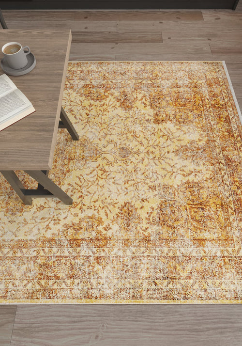 Bild 1 in Galerieansicht laden. The Ornate Beauty - Washable Rug - JR206, featuring a rustic yellow-gold pattern, is displayed on a wooden floor beside a light wood table with coffee and an open book—ideal for high-traffic areas.
