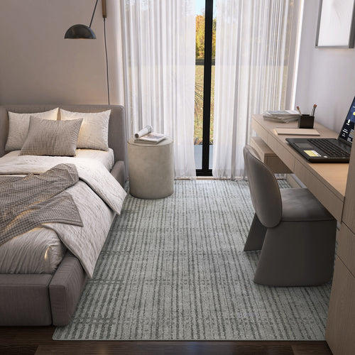 Load image 1 in gallery view. A modern bedroom features a gray upholstered bed, neutral bedding, an Ornate Hues - Washable Rug - JRN38 (Custom Size), a wooden desk with laptop and chair, a small round ottoman, and large windows with sheer curtains letting in natural light.