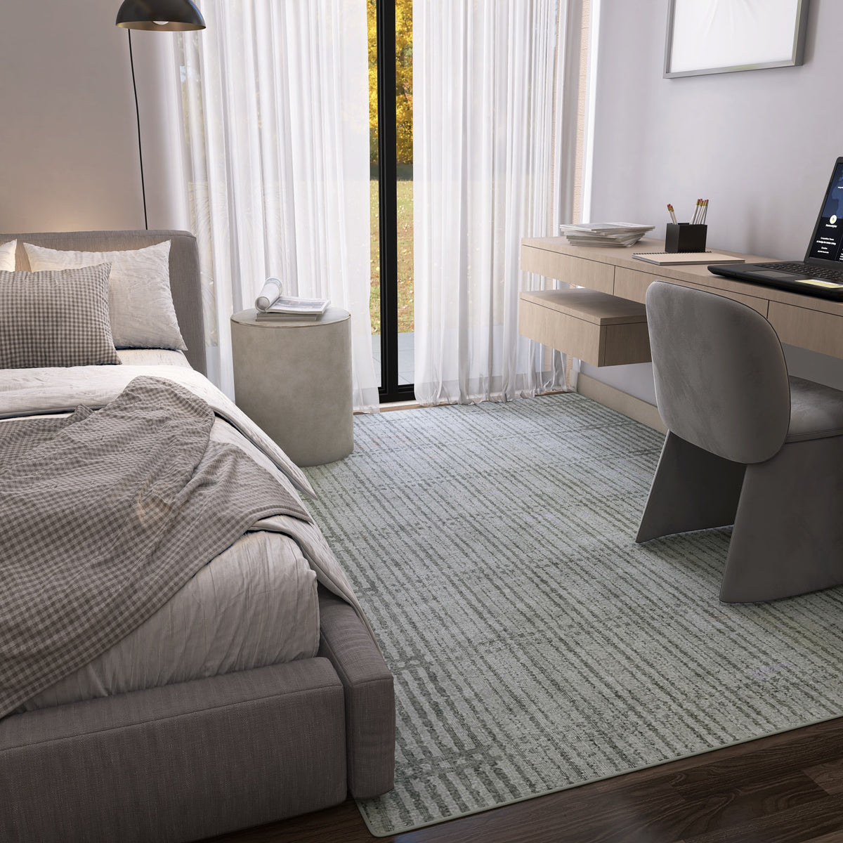 A modern bedroom features a large bed with neutral bedding, desk with computer, gray chair, round side table, and sheer curtains on tall windows. The Ornate Hues - Washable Rug - JRN38 (Custom Size) brings style and convenience to the floor.