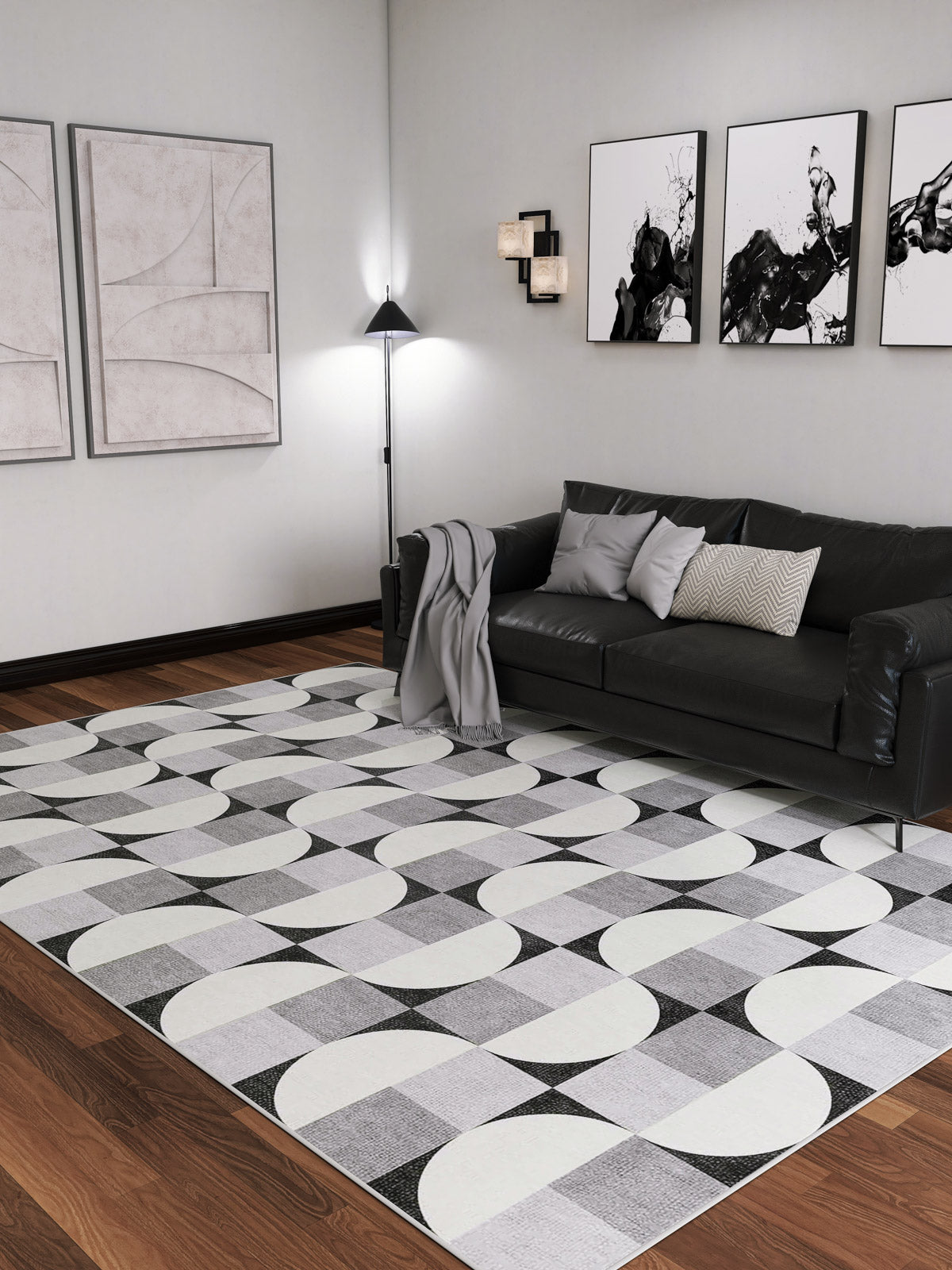 Modern living room with a black sofa, gray and white pillows, and a blanket. Abstract art decorates the walls, while the Patina Bloom - Washable Rug - JRN50 adds geometric style to the hardwood floor. Floor lamp and wall lights brighten the space.