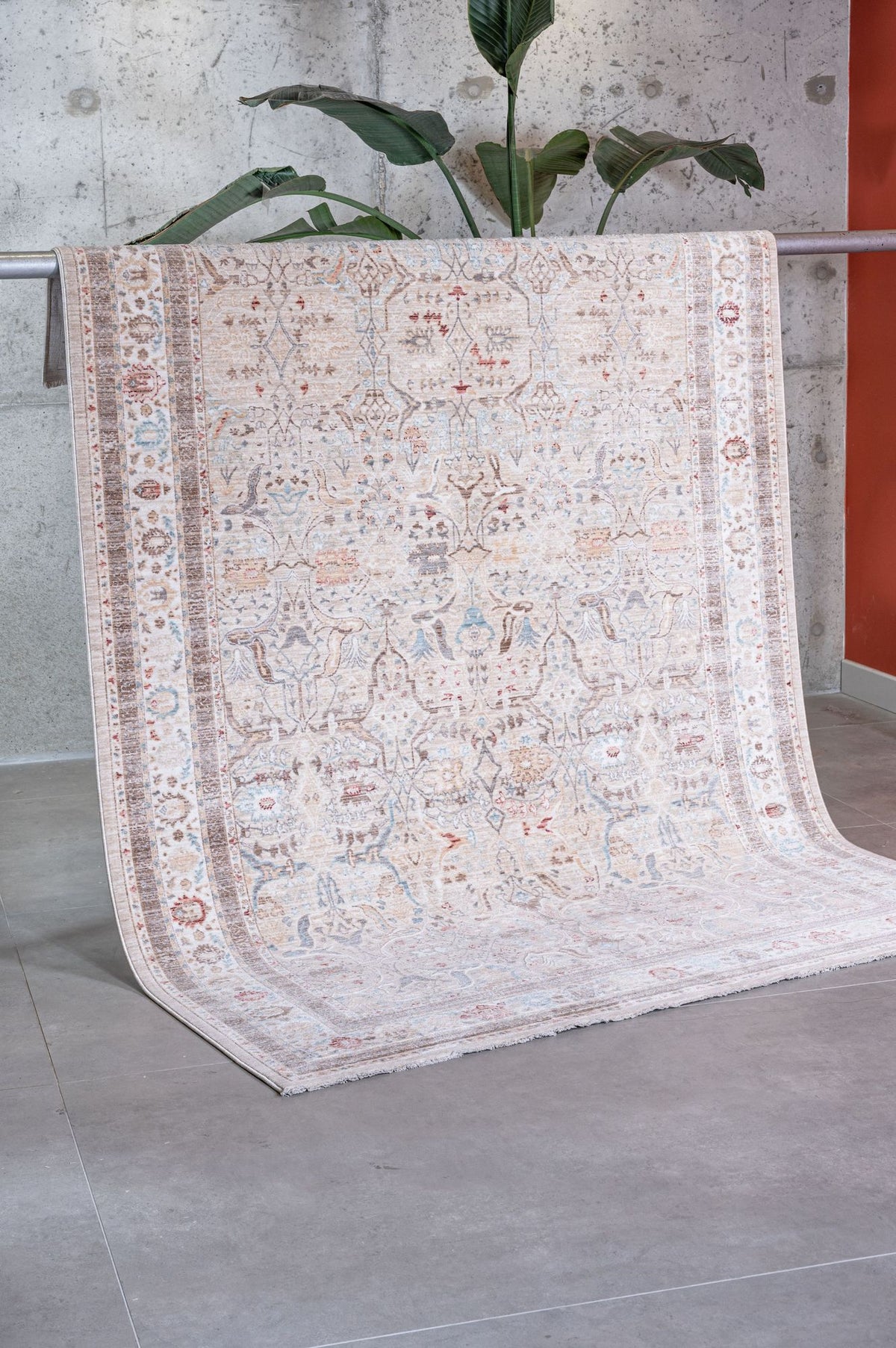 The Pearl Oushak - Area Rug - APX2931, featuring intricate beige vintage Persian designs and muted colors, is draped over a metal rail in a modern room with a concrete wall, gray tile flooring, and a tall green plant.