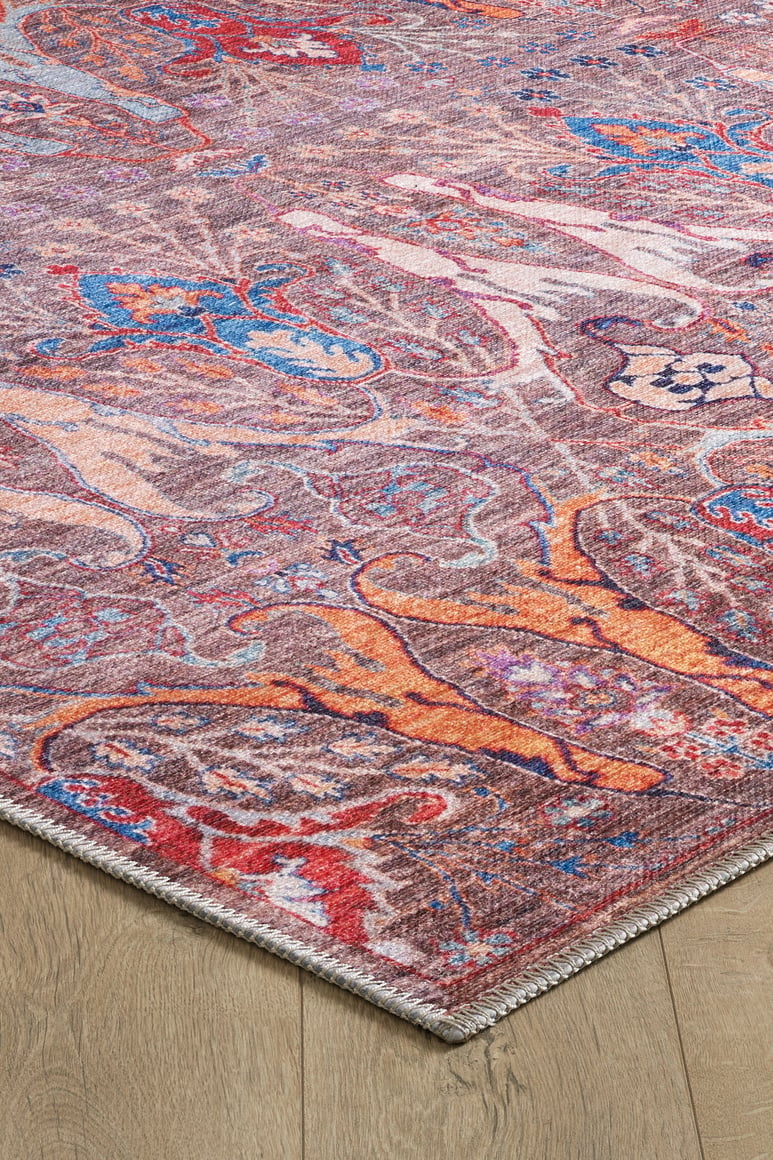 An angled close-up of the Peregrine Peachy Brown Washable Rug - LCC3022 shows its colorful abstract pattern in red, blue, orange, and purple on a light wood floor with detailed edge stitching.