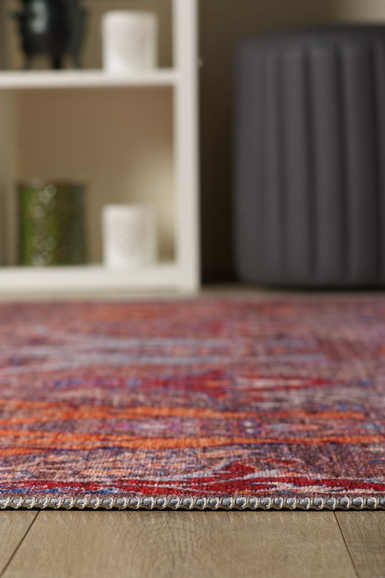 A close-up of the edge of the Peregrine Peachy Brown Washable Rug - LCC3022 (Outlet) US on a wooden floor, with a white shelving unit and gray ottoman softly blurred in the background.