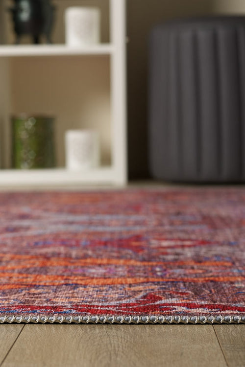 Load image 1 in gallery view. A close-up of the edge of the Peregrine Peachy Brown Washable Rug - LCC3022 (Outlet) US on a wooden floor, with a white shelving unit and gray ottoman softly blurred in the background.