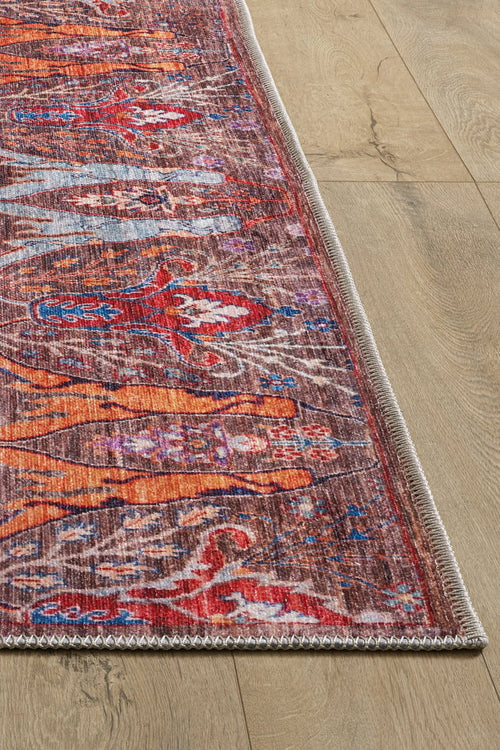Load image 1 in gallery view. A close-up of the Peregrine Peachy Brown Washable Rug - LCC3022 (Custom Size) shows its colorful orange, red, and blue designs with a stitched border on wood flooring—a vintage-inspired rug perfect for stylish, high-traffic areas.