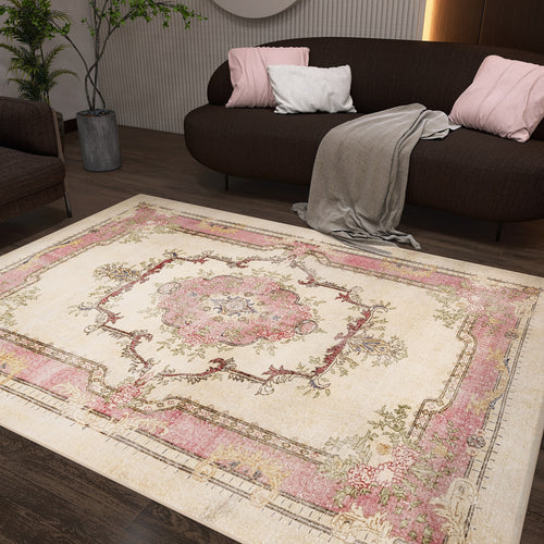 Load image 1 in gallery view. A living room with a dark brown sofa, pink and white pillows, a gray throw, a potted plant, and the Petal Mist - Washable Rug - JRS025 showcasing pink, cream, and floral patterns on a dark wood floor—ideal for modern spaces.