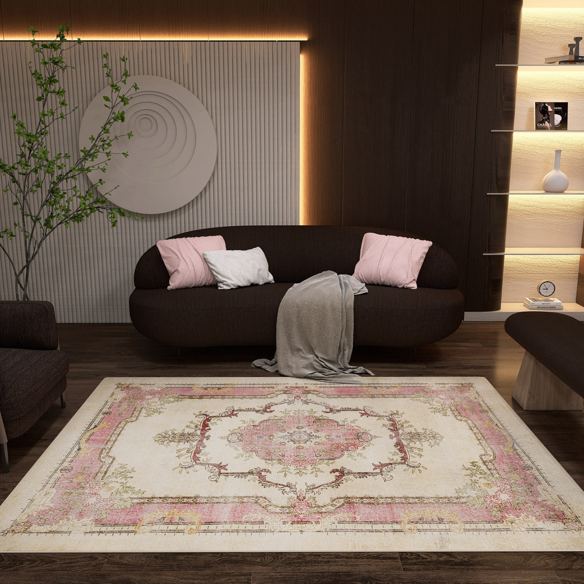 A cozy living room with a dark sofa, pink and white cushions, and a gray throw. The Petal Mist - Washable Rug - JRS025 with a pink and beige geometric pattern brightens the wooden floor. Shelves, soft lighting, wall art, and a plant complete the look.