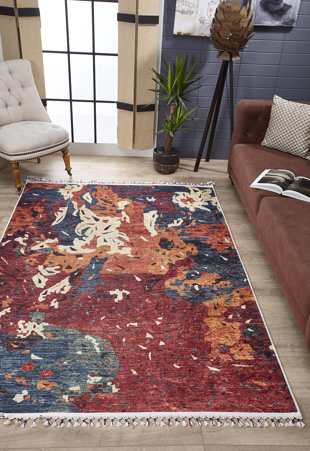 A modern living room with a Psychedelic Splash - Machine-Washable Rug (JR5096), abstract-patterned—ideal for allergy sufferers—brown sofa and pillow, cream tufted chair, potted plant, floor lamp, and large window with sheer curtains.