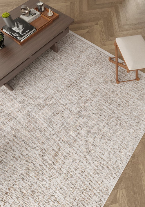 Load image 1 in gallery view. The Pure Bliss - Washable Rug - JR745, in light beige, sits on a herringbone wood floor beside a wooden coffee table with books, a tray, and a cup, plus a stool topped with a light cushion—ideal for high-traffic areas.