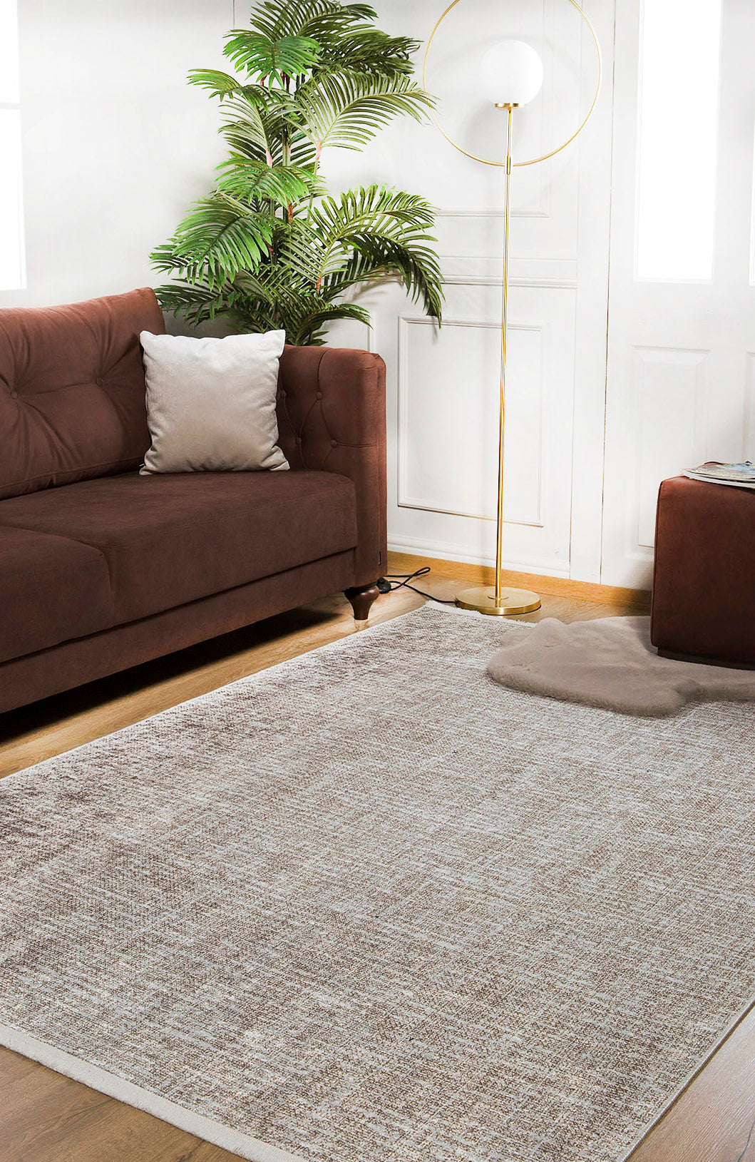 A cozy living room featuring a brown sofa, white pillow, Pure Bliss - Washable Rug - JR745 (Custom Size) ideal for allergy-sensitive homes, potted plant, floor lamp with round shade, and ottoman on wooden floors by white walls and doors.