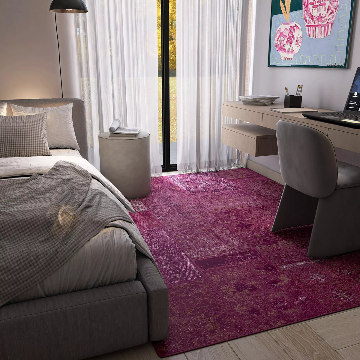 A cozy modern bedroom features a gray bed, rumpled bedding, and the Purple Mist - Washable Rug - JRN1038 in bright pink, perfect for high-traffic areas. Sunlight through sheer curtains highlights a desk with computer and gray chair.