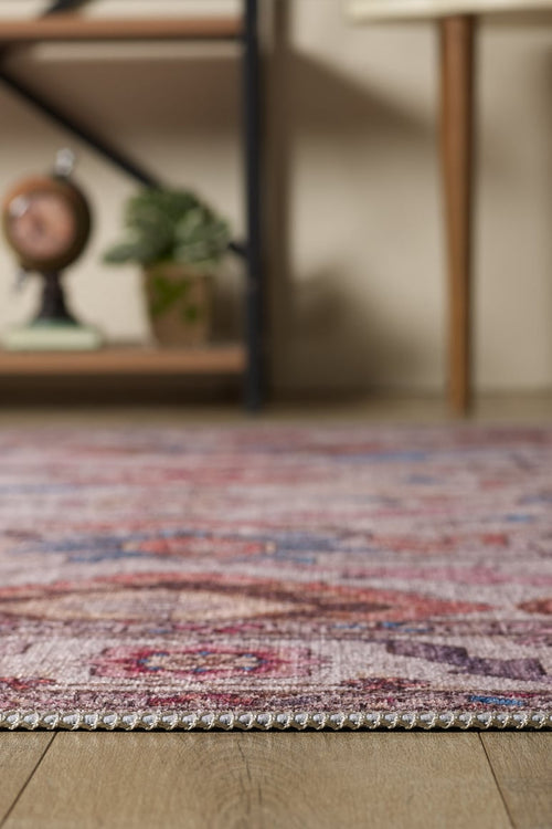 Load image 1 in gallery view. A close-up of the Quasar Traditional Washable Rug - LCC3024 (Custom Size) in vibrant pink, purple, and red tones on a wooden floor. Shelf, clock, and decor are blurred in the background—ideal for high-traffic areas.