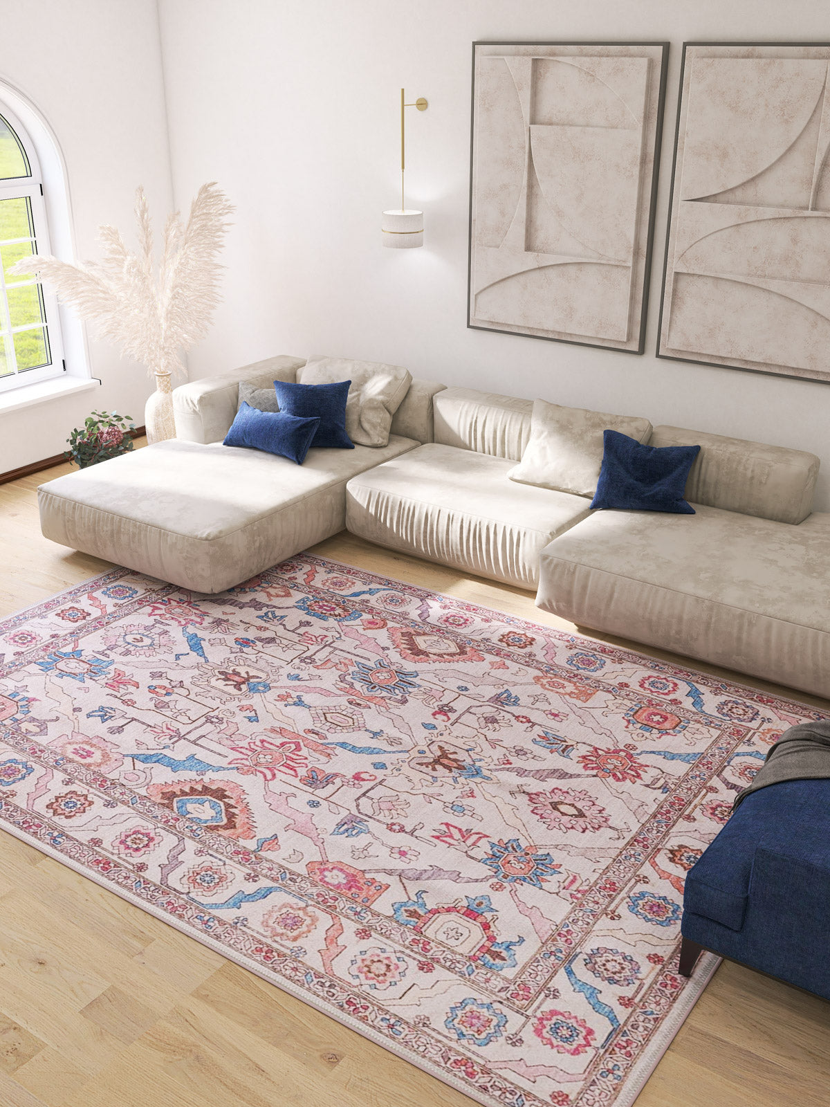 A modern living room with a large cream sectional, blue accent pillows, light wood floors, abstract wall art, natural light, and the Quasar Traditional Washable Rug - LCC3024 (Custom Size) in pink and blue—ideal for high-traffic areas.