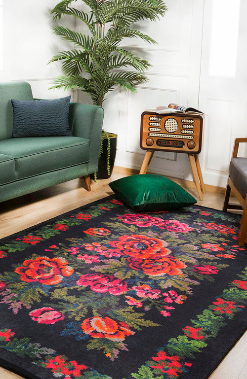 Load image 1 in gallery view. A living room features the Radiant Bloom - Washable Rug - JR294 (Custom Size) with bold red and pink florals on black, a green sofa, vintage radio on a wooden stand, potted plant, and a green velvet pillow on the floor.