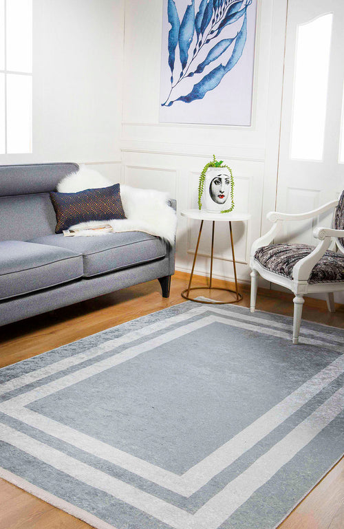 Load image 1 in gallery view. A modern living room features a gray sofa, patterned armchair, and the Refined Edge - Washable Rug - JR573 (Custom Size), ideal for high-traffic areas. A small round table with a face vase sits beneath blue leaf artwork on the white wall.