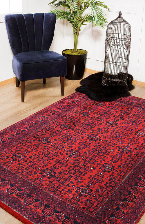 Load image 1 in gallery view. The Regal Accents - Washable Rug - JR36 (Custom Size) in a red pattern covers a wooden floor near a navy velvet chair, green potted plant, black fur rug, and an empty birdcage—ideal for high-traffic areas.