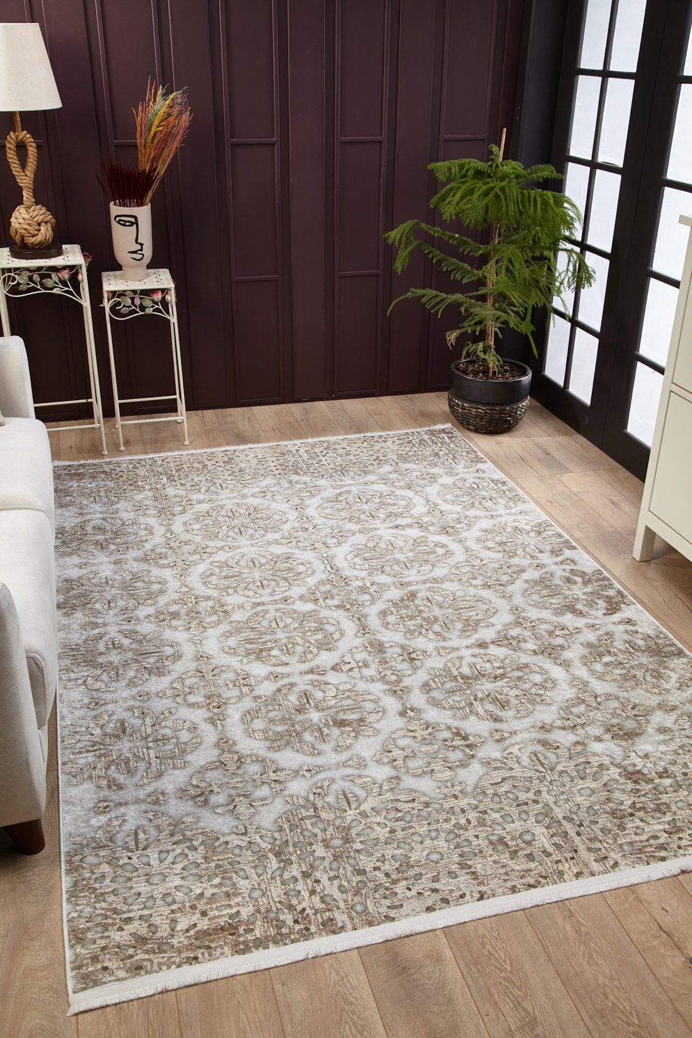 A living room with the Regal Embellishment - Washable Rug - JR1590 (Custom Size) in gray-anthracite on a wooden floor, white sofa, side table, decorative vase, potted plant, and dark paneled wall with tall window—ideal for high-traffic areas.