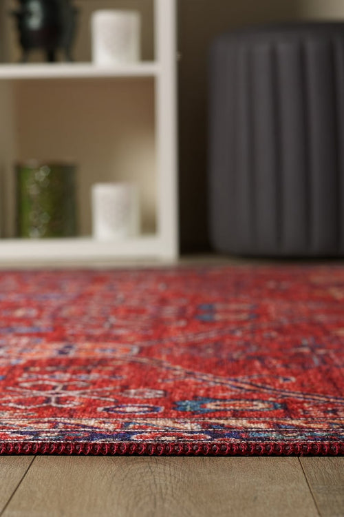 Load image 1 in gallery view. A close-up of the Rhapsody Authentic Red Washable Rug - LCC3033 (Outlet) US with a stylish pattern on a wooden floor; in the background, a white shelving unit holds candles and a green glass jar beside a dark gray ottoman—great for high-traffic areas.