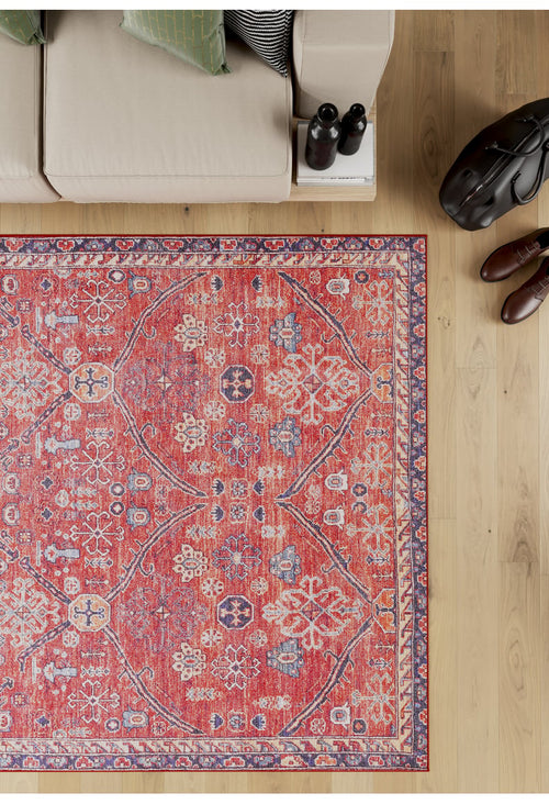 Load image 1 in gallery view. The Rhapsody Authentic Red Washable Rug (LCC3033) features blue and white accents and rests on a light wood floor near a beige sofa with green pillows, making it ideal for high-traffic areas. A side table holds two bottles, a black bag, and brown shoes.