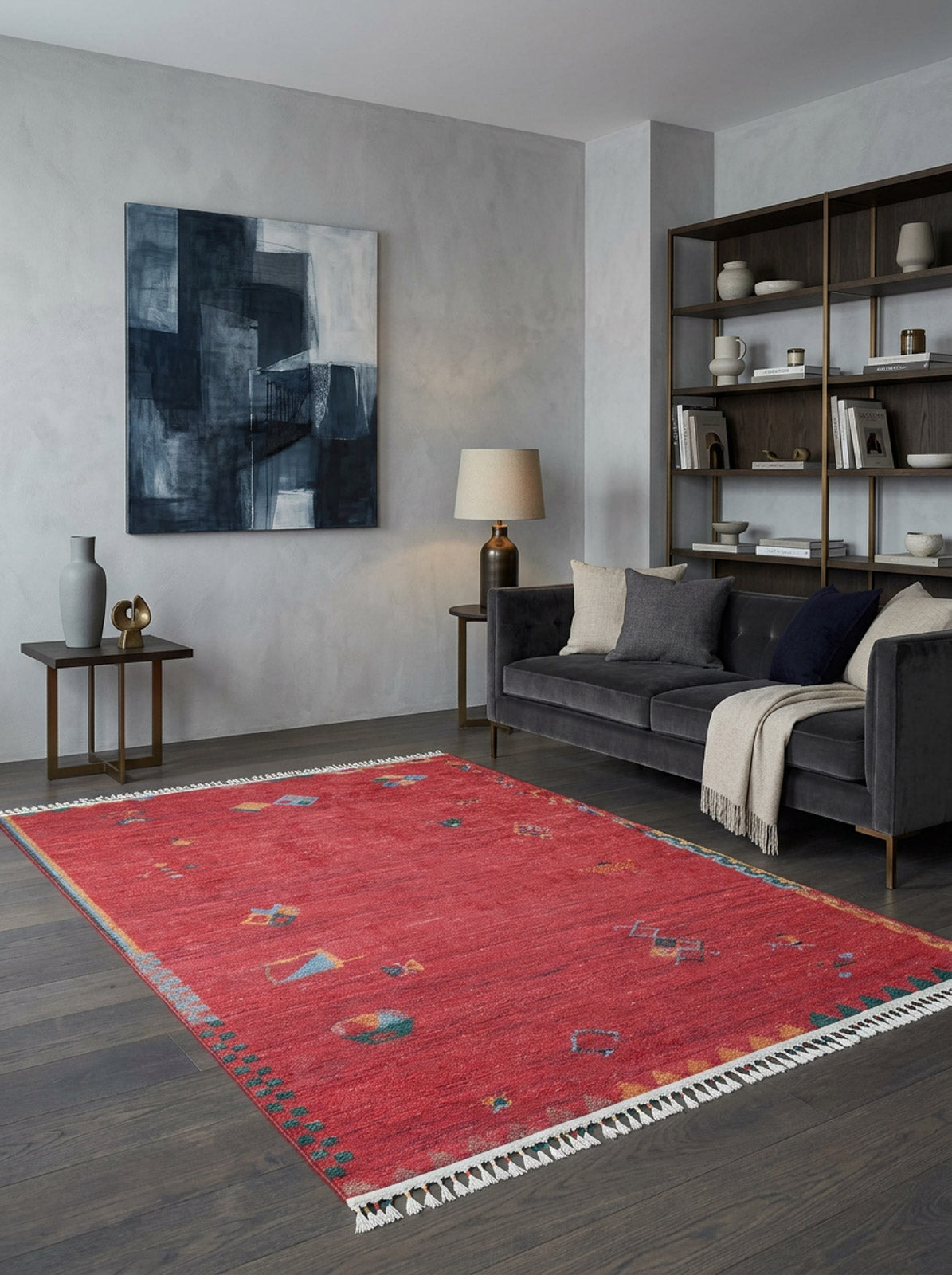 A modern living room featuring dark wood floors, the Rosy Blush Machine-Washable Rug (JRS5067) in red with patterns—ideal for high-traffic areas—a dark gray sofa, bookshelf, side table with lamp, vase, and an abstract blue and white painting on the wall.