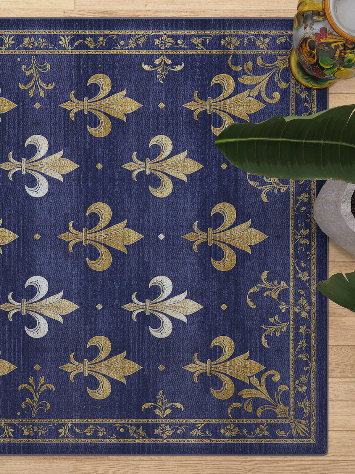 The Royal Fleur - Washable Rug - JRS016 features blue tones with gold and silver fleur-de-lis patterns and ornate borders, perfect for high-traffic areas. Styled on a light wood floor beside a leafy green plant and a small table.