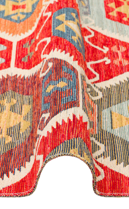 Load image 1 in gallery view. A close-up of the Ruby Grain - Kilim - NMD06 rug, featuring red, yellow, blue, and cream tribal geometric patterns, shown with a wavy folded edge to highlight its texture and thickness.
