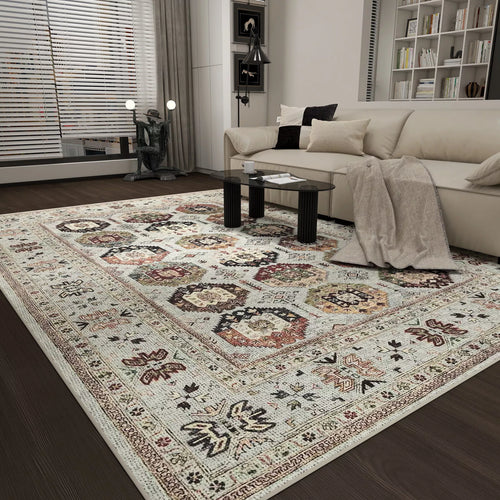 Load image 1 in gallery view. A modern living room with a beige sofa, glass coffee table, and the Rust Mosaic - Washable Rug - JRN1036, showcasing intricate geometric and floral designs in cream, brown, and red. Shelving and large windows complete the scene.