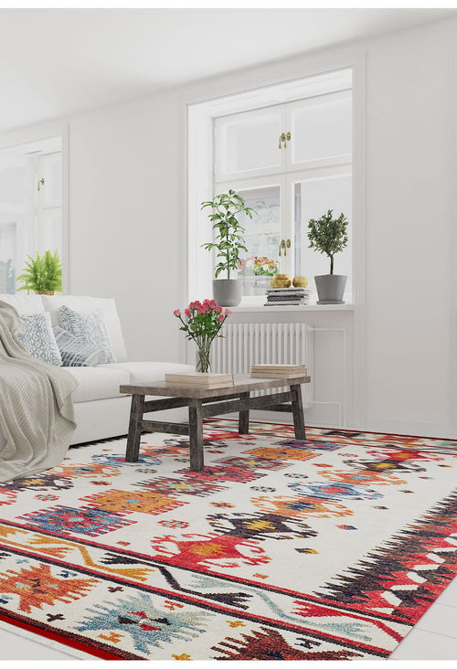 Load image 1 in gallery view. A bright, modern living room with large windows, a white sofa with patterned cushions, a rustic wooden coffee table topped with flowers, plants on the windowsill, and the vibrant Rustic Refrain - Washable Rug - JR149 on the floor.