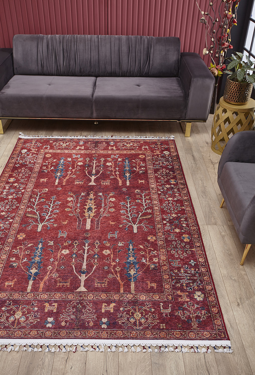 A living room features a dark gray sofa, matching armchair, and the Rustic Retreat - Washable Rug - JR5095 (Custom Size), with tree and animal designs—ideal for high-traffic areas. A plant in a stand sits by the sofa against a red-striped wall on light wood floors.