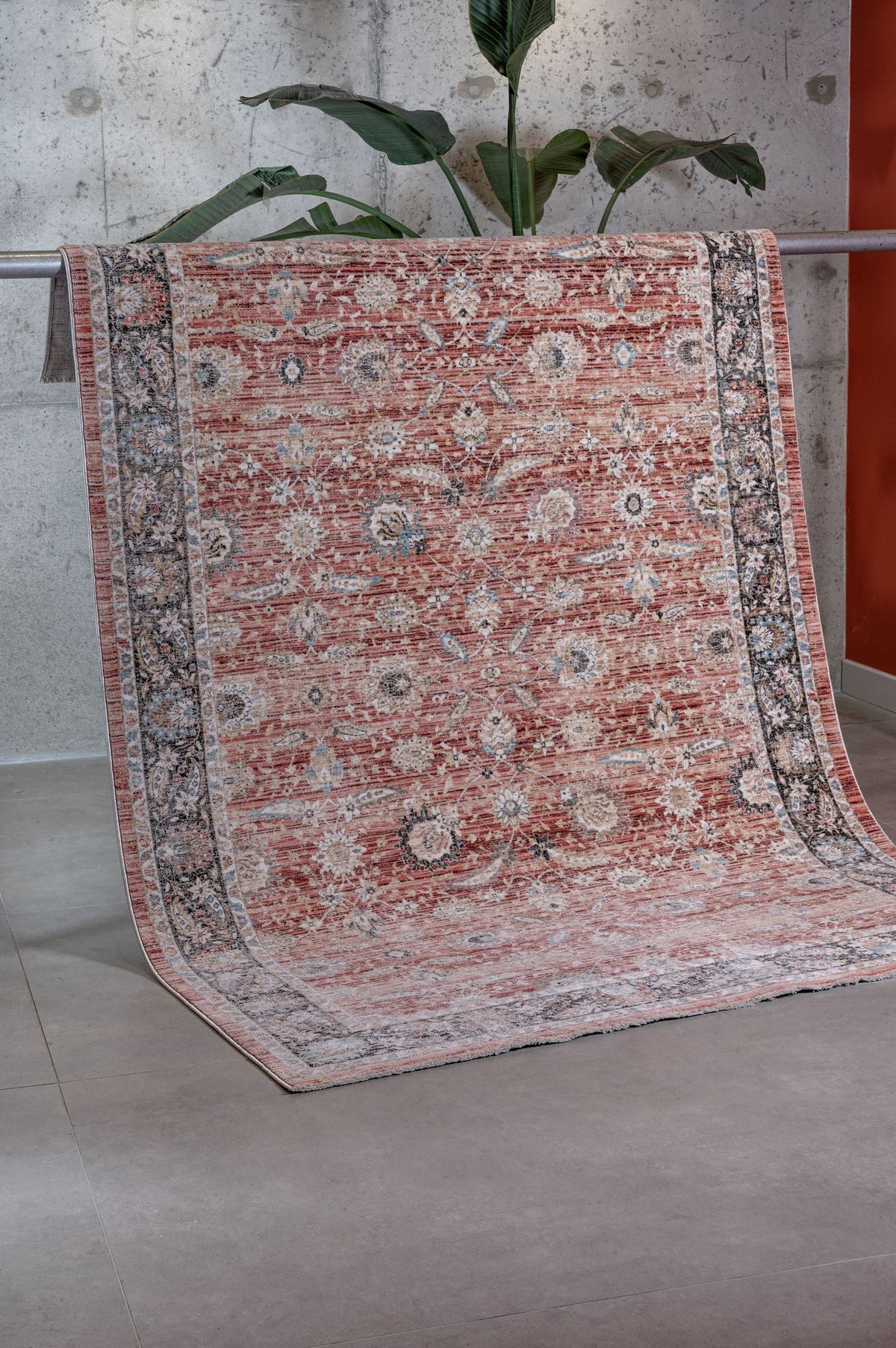 The Rustic Bloom - Area Rug - APX2961, featuring red and beige floral motifs, hangs over a metal railing in a modern room with a concrete wall, gray floor, and a green plant in the background.