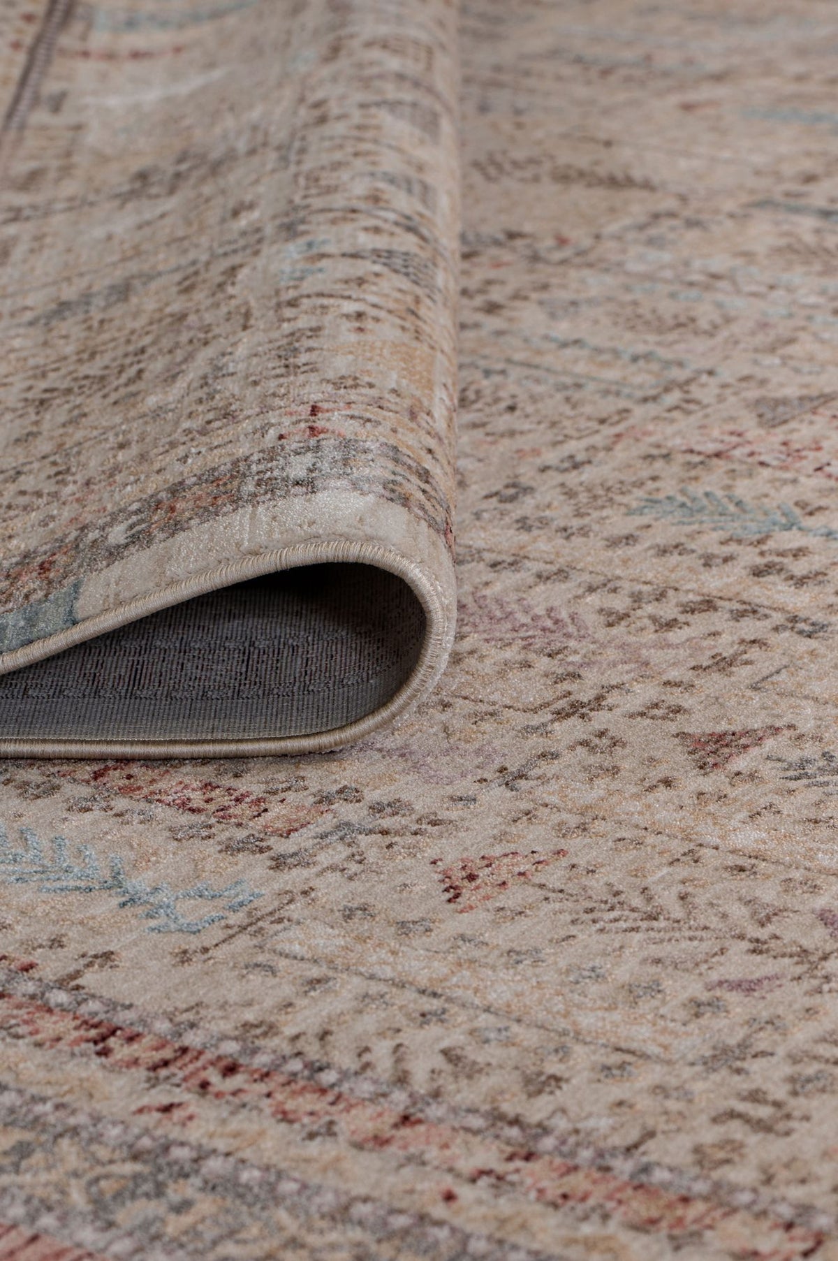 Beige area rug with a distressed traditional pattern in muted red, blue, and grey, shown with a corner folded over.