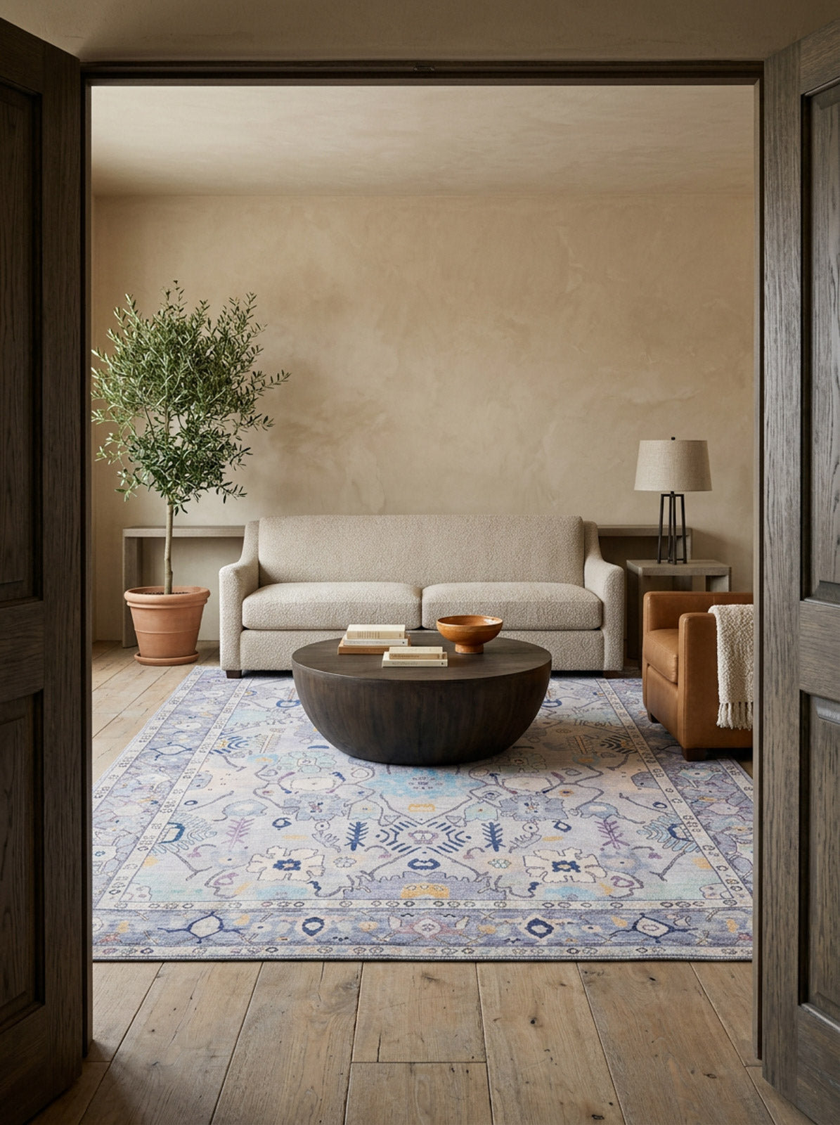A cozy living room features a light beige sofa, tan armchair, and round wooden table. The Seraphix Pastel Bordered Washable Rug (LCC3011) covers the wood floor, while neutral walls and a potted tree enhance the inviting vibe.