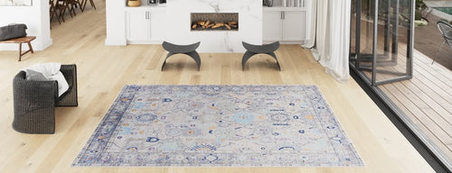 Load image 1 in gallery view. A modern living room with light wood floors, a Seraphix Pastel Bordered Washable Rug (LCC3011) in blue-gray hues, a black wicker chair, two curved stools, a fireplace, and large glass doors—perfect for high-traffic areas.