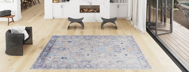 A spacious, modern living room with light wood floors, a Seraphix Pastel Bordered Washable Rug (LCC3011) ideal for high-traffic areas, a black woven chair, two gray stools by the fireplace, and floor-to-ceiling windows welcoming natural light.