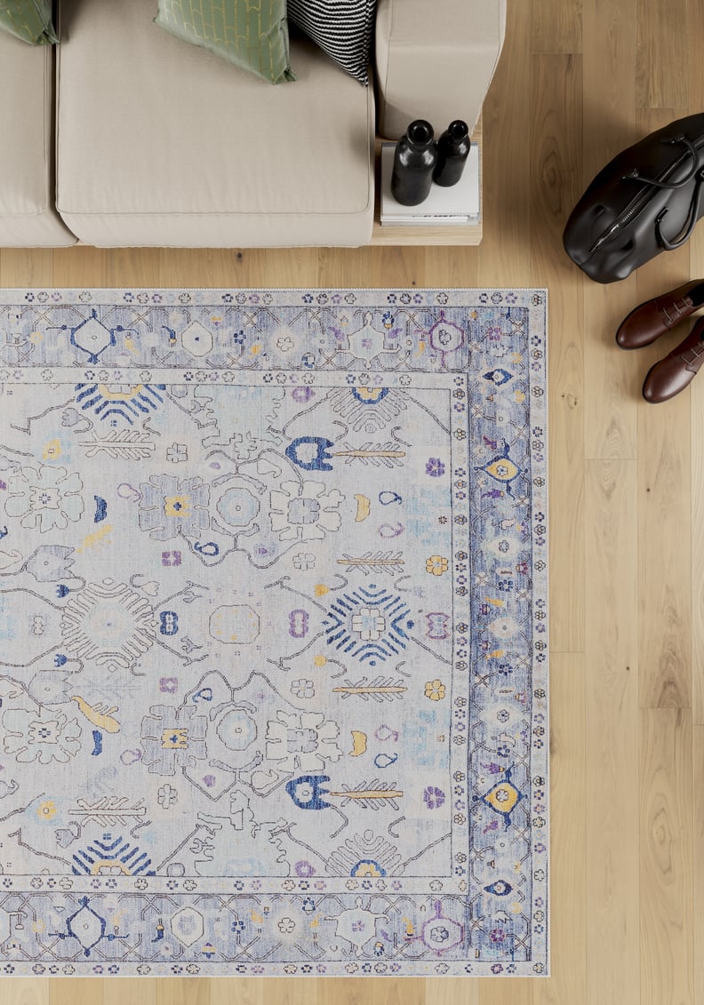A top-down view of the Seraphix Pastel Bordered Washable Rug - LCC3011 (Custom Size) in blue with yellow and beige designs on wood flooring, near a beige sofa with green pillows—ideal for high-traffic areas.