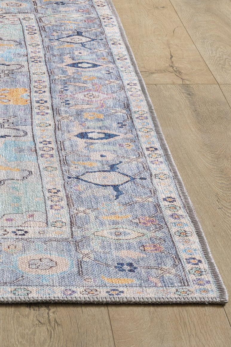 A close-up of the Seraphix Pastel Bordered Washable Rug - LCC3011 (Custom Size) shows its rectangular blue design with geometric and floral patterns on a light wood floor—ideal for high-traffic areas.