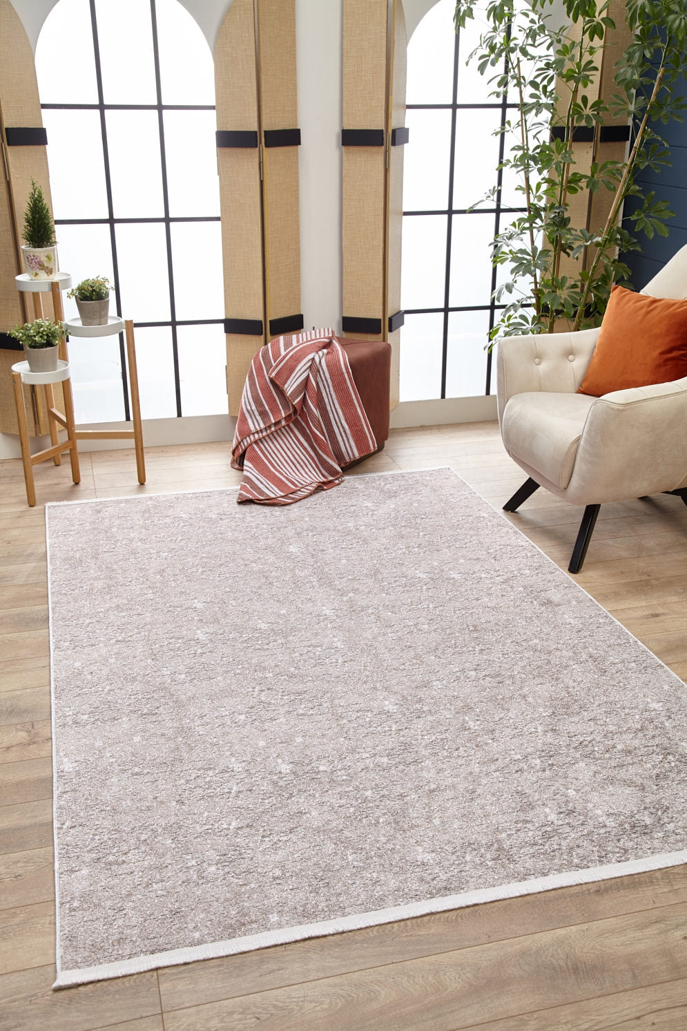 The Slate Charm - Washable Rug - JR1740 (Custom Size) in light gray sits on a wooden floor beside a beige armchair with an orange pillow, a brown ottoman with a striped blanket, and large arched windows—perfect for high-traffic areas.