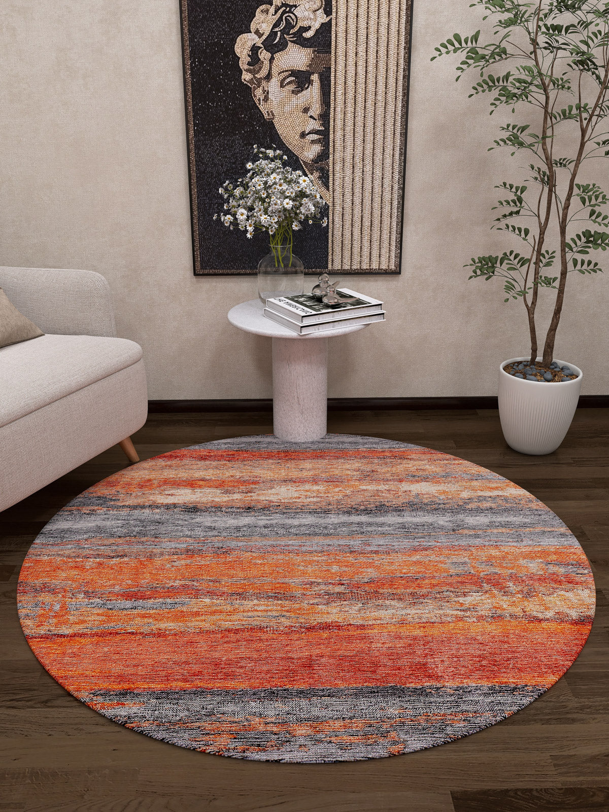 A modern living room features the Sleek Brushed - Washable Rug - JR431 (Custom Size) in orange and gray, ideal for high-traffic areas, along with a small white side table holding books and flowers, a potted plant, a beige sofa, and classical sculpture wall art.