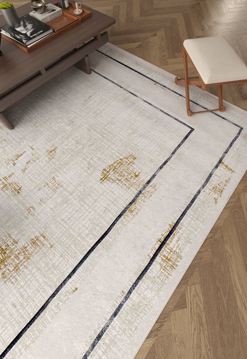 A modern living room with the Sleek and Contemporary - Washable Rug - JR1365 (Outlet) US, a flat weave rug in cream and beige with golden accents and dark lines, on a herringbone wood floor, plus a wooden coffee table and cushioned stool.