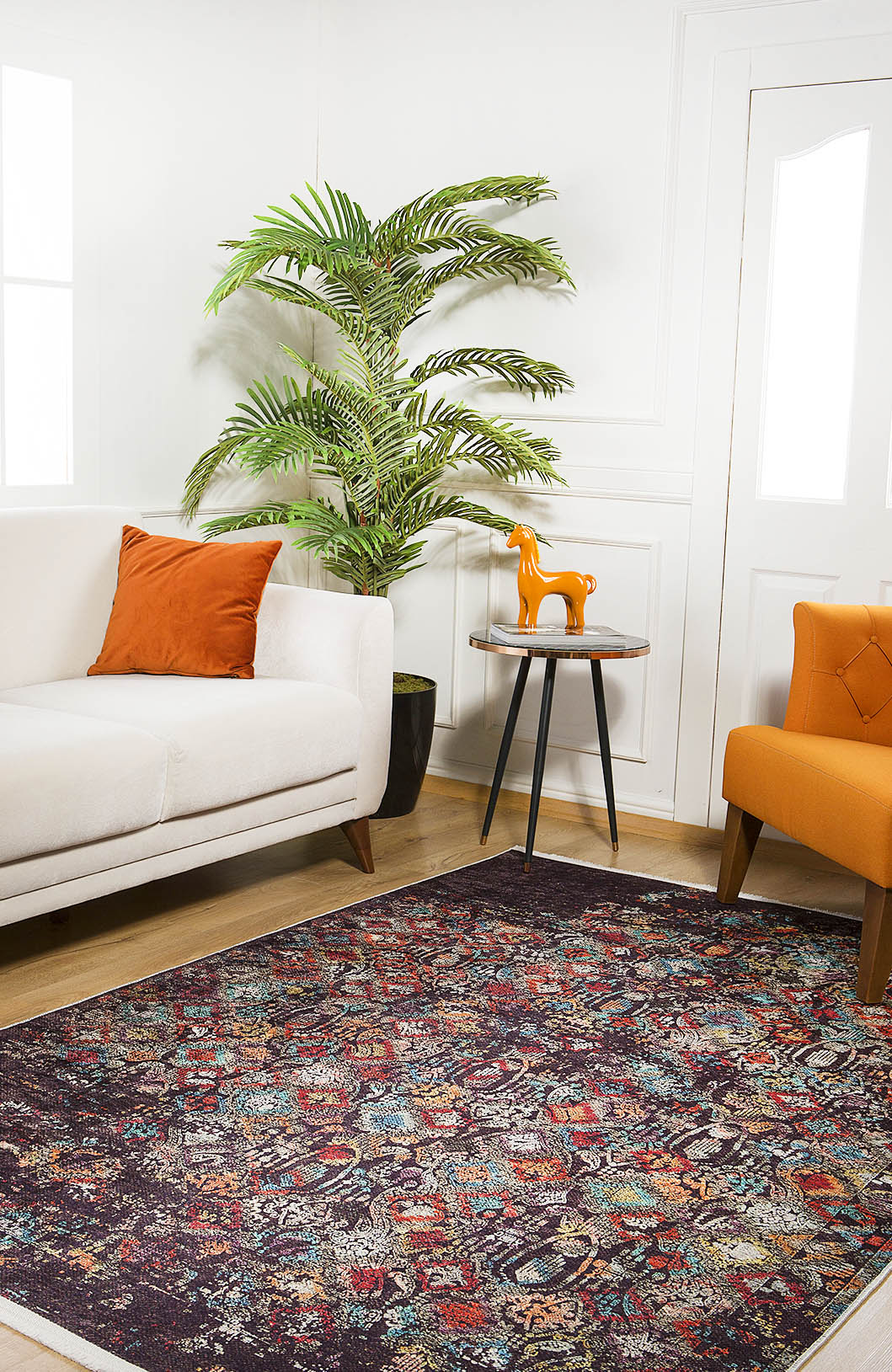 A living room featuring the Soft Haze - Washable Rug - JR1094 (Custom Size) for high-traffic areas, a white sofa with an orange pillow, orange armchair, small round table with an orange horse figurine, potted plant, and white walls.