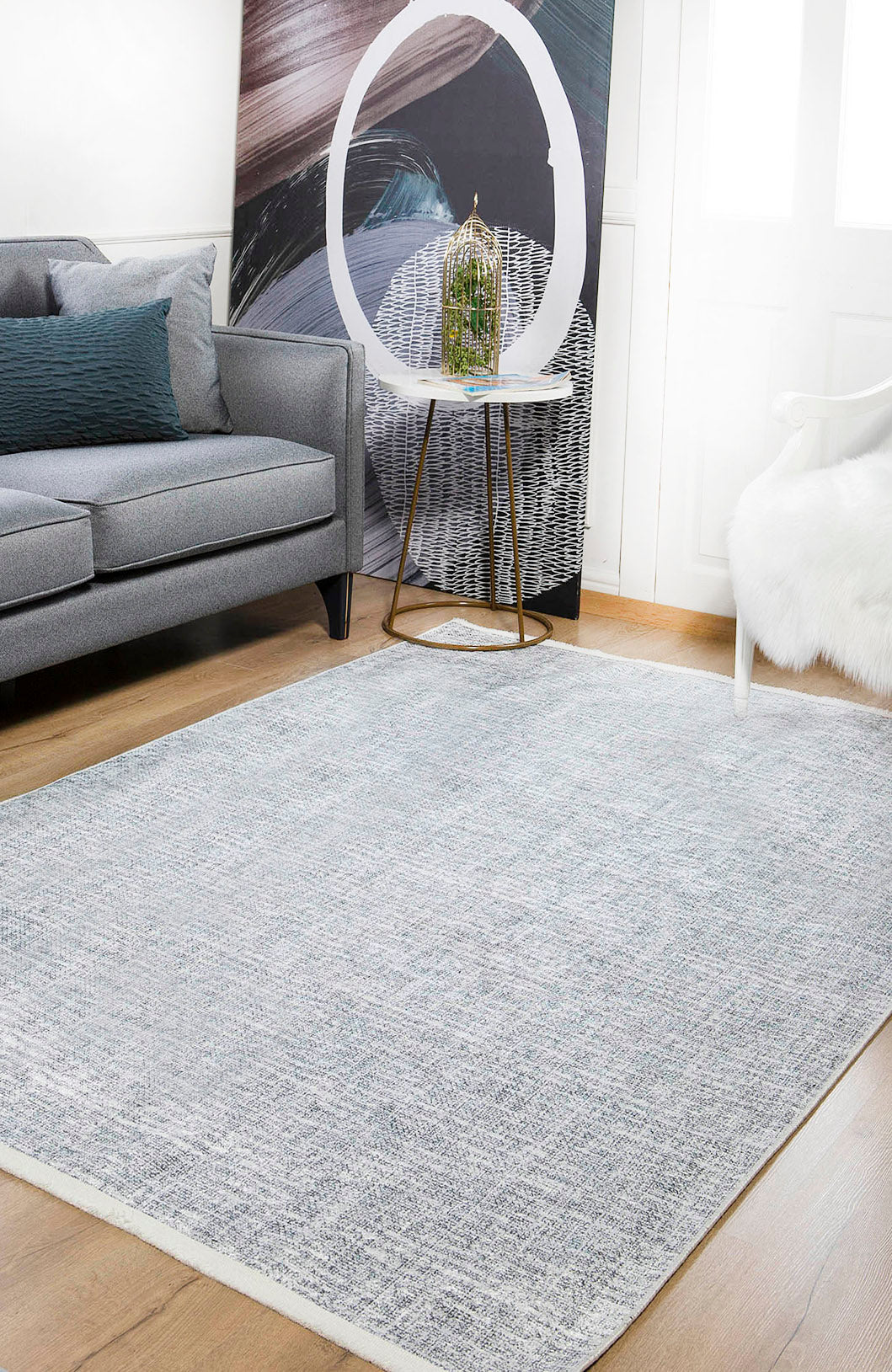 A modern living room with a gray sofa, the Softly Chic - Washable Rug - JR747 in blue-gray for high-traffic areas, a small side table with a glass birdcage, abstract wall art, a white fur chair, and light wood flooring.