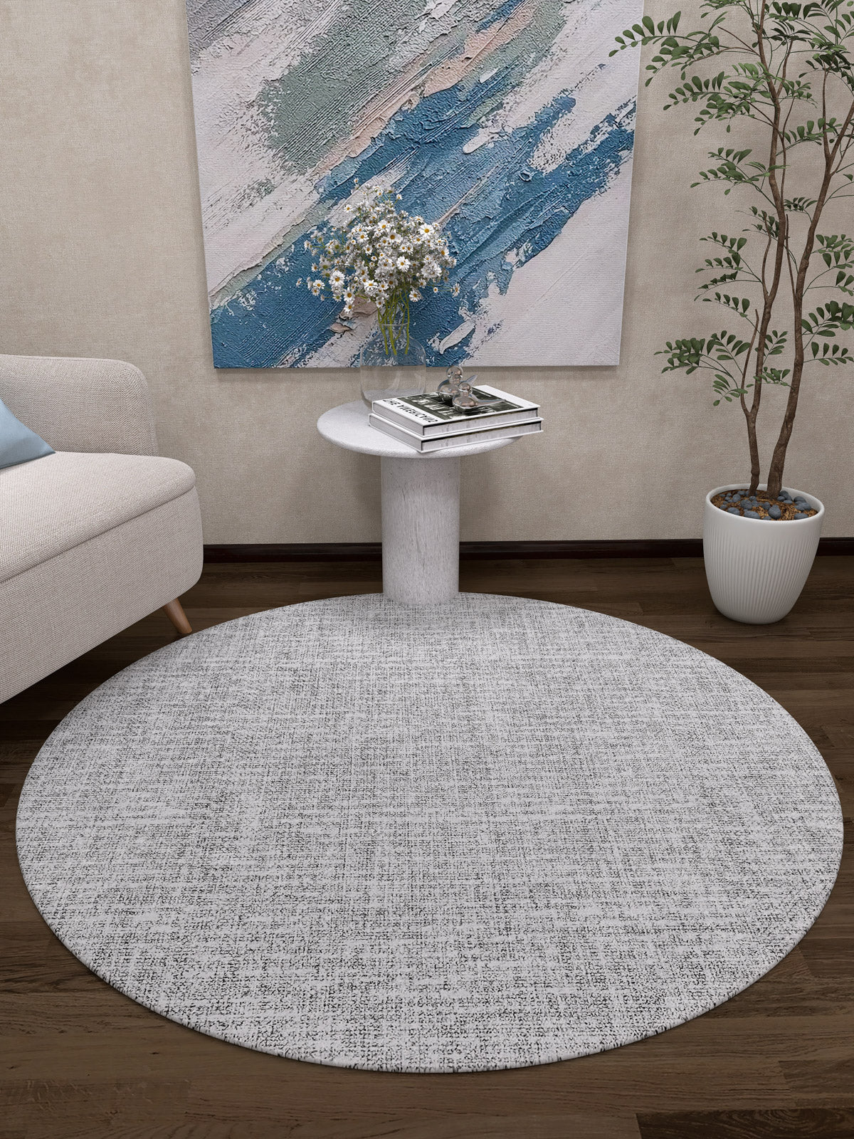A modern living room corner featuring the Softly Chic - Washable Rug - JR747 in gray-anthracite, a round white side table with books and white flowers, a potted plant, beige sofa, and abstract painting—perfect for high-traffic areas.